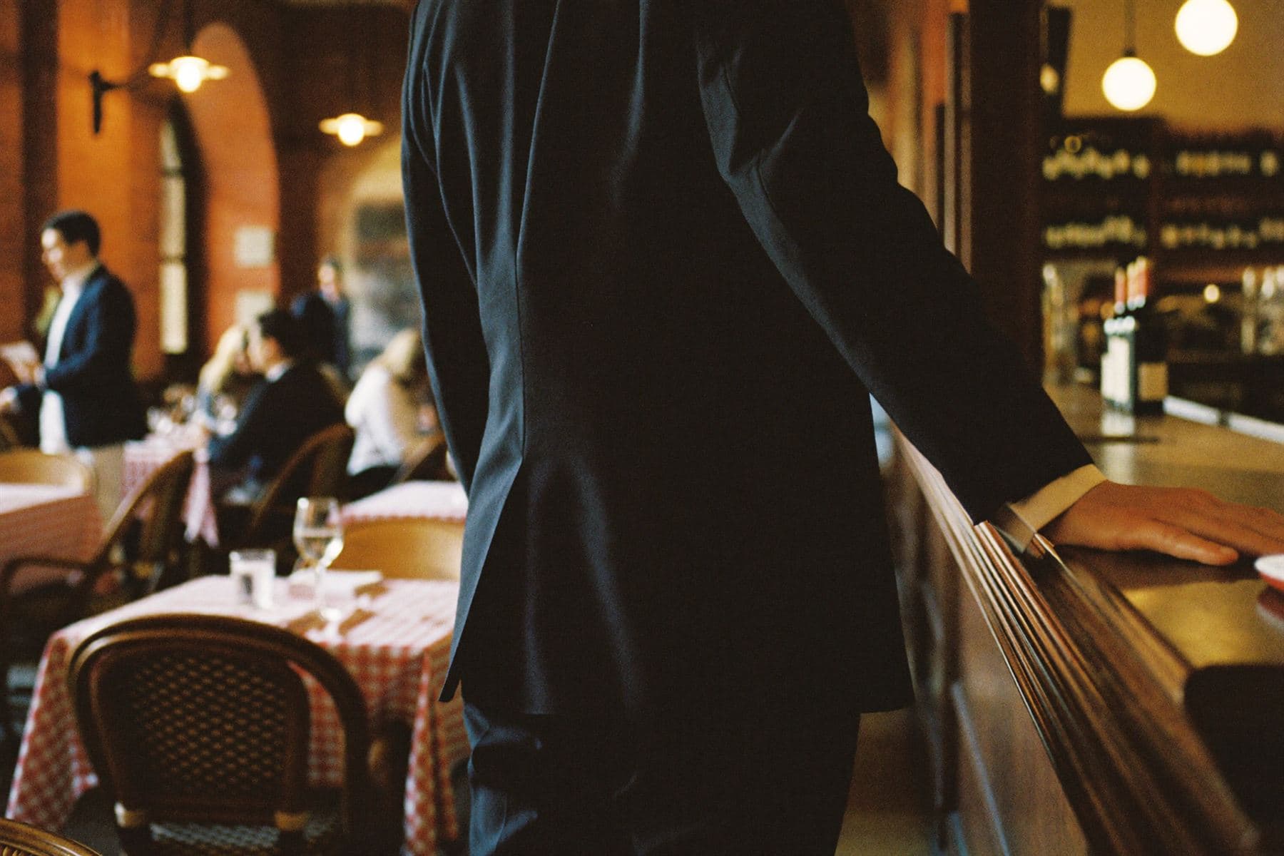 Una persona de espaldas, apoyada en la barra, observa el salón de una trattoria durante el servicio.