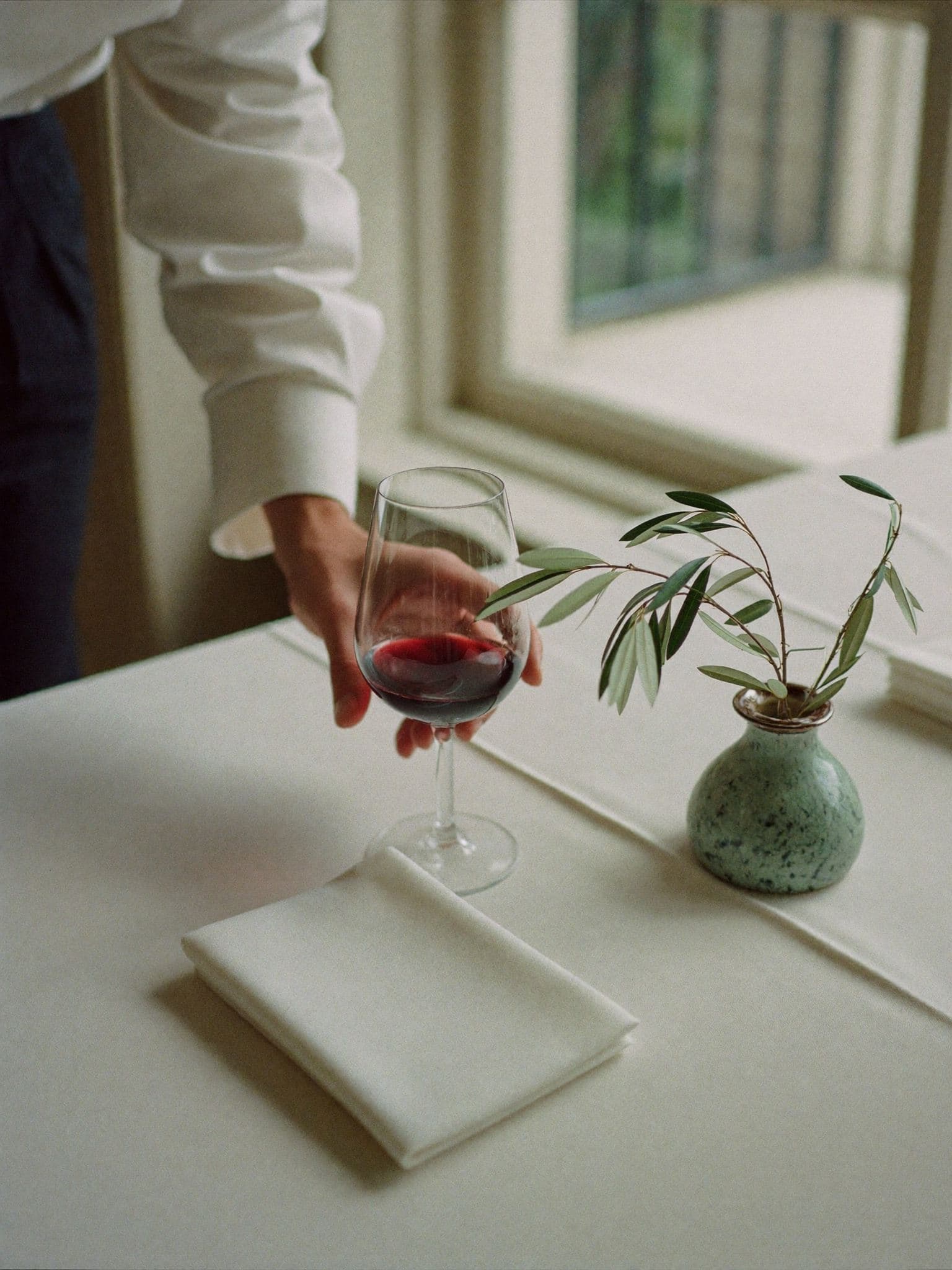 Un camarero apoya una copa de Malbec sobre un mantel blanco junto a una rama de olivo.